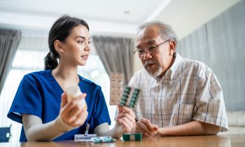 Asian Senior elderly male patient consult with physician nurse at nursing home care. Caregiver therapist pharmacist girl hold medicine pills bottle, explain prescription to older disabled guy in house