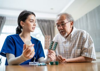 Asian Senior elderly male patient consult with physician nurse at nursing home care. Caregiver therapist pharmacist girl hold medicine pills bottle, explain prescription to older disabled guy in house