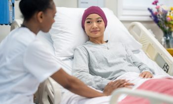 Oncology Rounds A female nurse of African decent sits at the bedside of a female patient as she checks in on her during rounds.  The patient is dressed comfortably and has a head scarf on to keep warm.