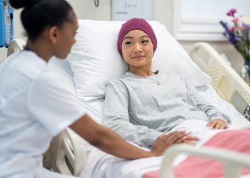 A female nurse of African decent sits at the bedside of a female patient as she checks in on her during rounds.  The patient is dressed comfortably and has a head scarf on to keep warm.