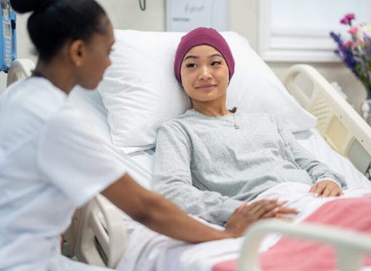Oncology Rounds A female nurse of African decent sits at the bedside of a female patient as she checks in on her during rounds.  The patient is dressed comfortably and has a head scarf on to keep warm.